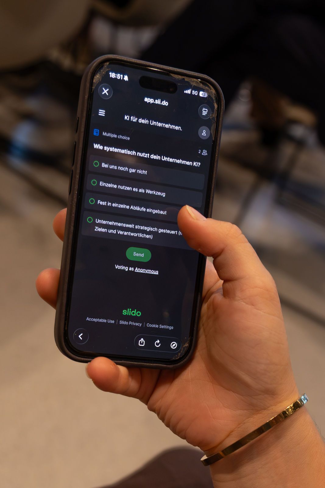 Eine Hand hält ein Smartphone mit der geöffneten Slido-Abstimmung und den vier Antwortmöglichkeiten der zweiten Frage.