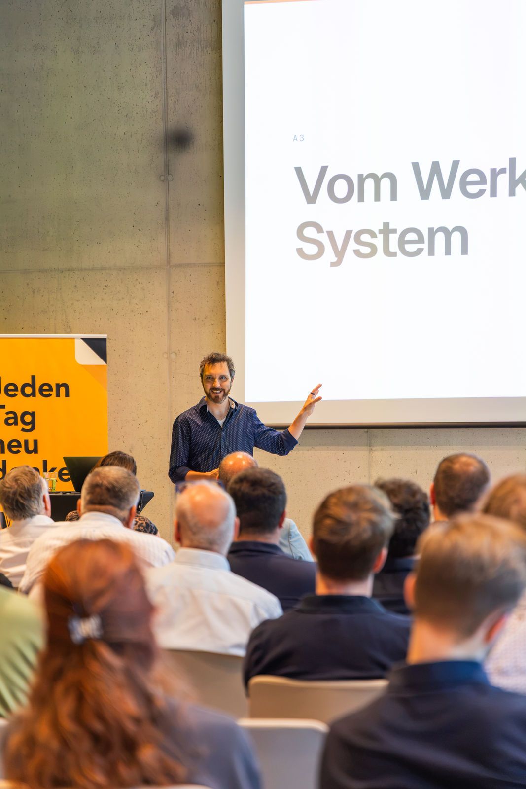 Der Vortragende zeigt auf die Leinwand, auf der die Überschrift „Vom Werkzeug zum System“ steht.